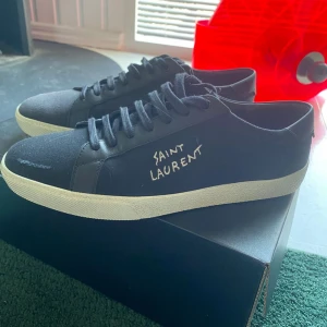 Svarta Saint Laurent sneakers - Snygga svarta sneakers från Saint Laurent med vit broderad logga på sidan. Skorna har snörning, vit platt sula och är gjorda i en mix av canvas och skinn. Perfekta för dig som gillar stilrena och exklusiva sneakers med en cool touch.
