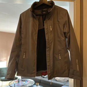 Grå vindjacka från Helly Hansen - Snygg grå vindjacka från Helly Hansen med hög krage, dragkedja och flera praktiska fickor. Jackan har Helly Tech Protection-tyg och HH-loggor på bröst, ärm och rygg. Perfekt för blåsiga dagar och outdoor-aktiviteter.