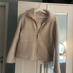Beige teddyjacka med dragkedja - Mysig beige teddyjacka med krage och två stora fickor framtill. Jackan har en hel dragkedja och är tillverkad i mjukt fleece-material. Perfekt för att hålla sig varm och samtidigt se chill ut. Passar dig som gillar en enkel och clean stil.