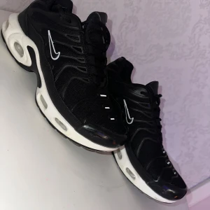 Nike Air Max Plus svarta sneakers - Säljer ett par svarta Nike Air Max Plus sneakers med vit sula och tydliga luftbubblor. Skorna har en sportig vibe med mesh och syntetmaterial, svarta snören och vita detaljer på sidorna samt Nike-logga. Perfekta för dig som gillar streetstyle och bekväma skor.