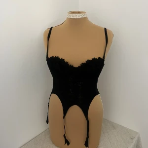 Svart spetskorsett med blommönster - Säljer en svart korsett med blommig spets framtill och justerbara axelband. Modellen har genomskinlig mesh på ryggen och hakknäppning. Utskurna detaljer och hängande strumpeband ger en edgy vibe. Perfekt för dig som vill ha något extra till outfiten.