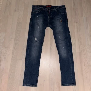Dsquared2 blå jeans med slitningar - Snygga blå jeans från Dsquared2 med slitna detaljer och broderad lönnlöv på bakfickan. Klassisk femficksmodell med normal passform och diskret tvättad look. Röd Dsquared2-logga på insidan och svart patch bak i midjan. Perfekta för dig som gillar streetstyle.