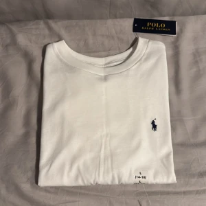 Vit t-shirt från Polo Ralph Lauren - Vit t-shirt från Polo Ralph Lauren med klassisk rund hals och den ikoniska broderade loggan i marinblått på bröstet. Tillverkad i mjuk bomull för skön komfort. Perfekt för dig som gillar stilrena och tidlösa plagg med premiumkänsla.