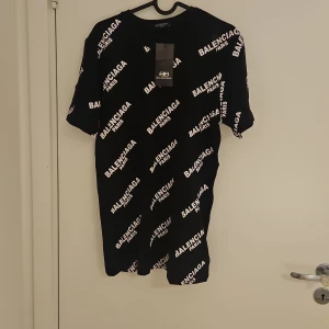 Svart t-shirt med logga - Svart t-shirt med vit logotyp tryckt över hela plagget. Klassisk rund hals och korta ärmar. T-shirten är gjord i mjuk bomull och har en loose passform som ger en chill vibe. Perfekt för dig som gillar streetwear och vill sticka ut med designerstil.