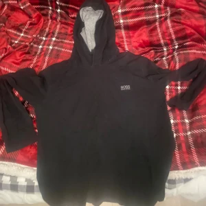 Svart hoodie från Hugo Boss - Svart hoodie från Hugo Boss med grått foder i huvan och diskret logga på bröstet. Tröjan har lång ärm och klassisk huva, perfekt för chill dagar. Materialet känns mjukt och skönt mot huden. Snygg och enkel design som funkar till jeans eller joggers.