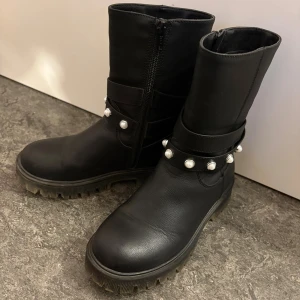 Svarta boots med pärldetaljer - Snygga svarta boots i skinnimitation med grov sula och dragkedja på sidan. Insidan är fodrad med mjukt fuskpälsfoder för extra värme. Dekorativa remmar med pärldetaljer runt ankeln ger en cool touch. Perfekta för kalla dagar och passar till många outfits.