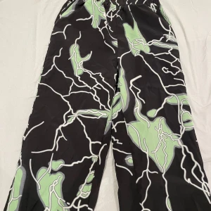 Svarta byxor med grön abstrakt print - Säljer ett par svarta byxor med unikt abstrakt mönster i grönt och vitt. Byxorna har en loose passform och elastisk midja för extra komfort. Perfekta för dig som vill sticka ut med en cool och modern stil.