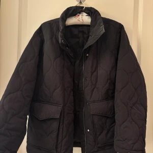 Svart quiltad jacka från & Other Stories - Quiltad svart jacka från & Other Stories med hög krage och stora fickor framtill. Jackan har ett vågigt quiltat mönster och stängs med både dragkedja och tryckknappar. Hör av er om ni har några funderingar!💗