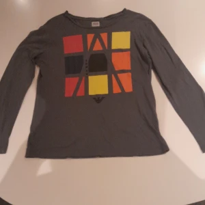 Grå långärmad tröja Armani Junior - Grå långärmad tröja från Armani Junior med färgglatt grafiskt tryck i rött, gult, orange och svart på bröstet. Mjuk bomullskänsla och rund halsringning. Perfekt för dig som gillar stilrena men ändå unika plagg.
