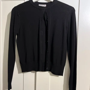 Svart kofta med knappar - En klassisk svart kofta med rund halsringning och knäppning framtill. Långärmad och i ett mjukt, tunt material som passar perfekt till lager-på-lager. Enkel och stilren design som funkar till det mesta.