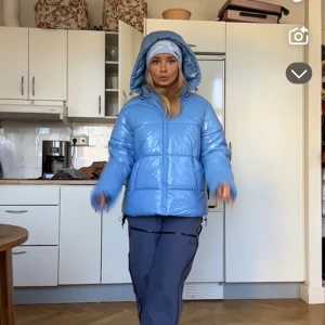 Blå pufferjacka med huva - Säljer en glansig ljusblå pufferjacka med huva och dragkedja. Jackan är vadderad och har en oversized vibe, perfekt för kalla dagar. Snygg och trendig modell som håller dig varm hela vintern.