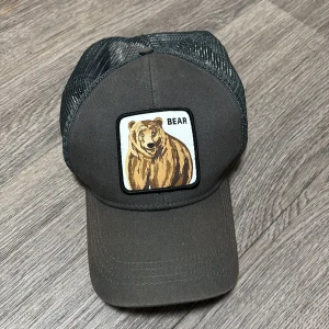 Svart keps LC Waikiki med björnpatch - Säljer en svart truckerkeps från LC Waikiki med mesh på sidorna och bak. Framtill finns en broderad patch med en björn och texten 'BEAR'. Kepsen har böjd skärm och justerbar passform. Perfekt för dig som gillar djurmotiv och streetstyle.