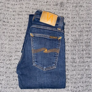 Nudie Jeans - Snygga mörkblå jeans från Nudie Jeans med klassiska orange sömmar och logga på bakfickan. Modellen har en slim passform och är perfekta för dig som gillar stilrena jeans. Höftmåttet på de är 31cm och längdmåttet på de är 87cm. Tveka inte på att ställa frågor!