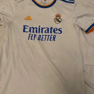 Real Madrid vit fotbollströja Adidas - Säljer en officiell Real Madrid fotbollströja från Adidas i vit med blå och orange detaljer. Tröjan har korta ärmar, klubbmärke på bröstet och Emirates Fly Better-tryck framtill. Tillverkad i lätt och ventilerande material, perfekt för match eller träning.
