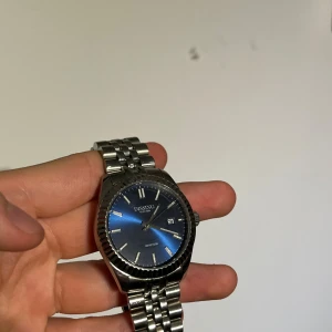 Dissing klocka med blå urtavla - Stilren klocka från Dissing med blå urtavla och datumvisning. Boetten och länken är i silverfärgat metall och klockan kommer i original svart box. Klassisk design med räfflad bezel och tydliga indexmarkeringar. Man får med länkar så man kan göra den större om man vill de.