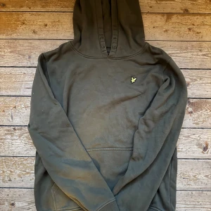 Mörkgrön hoodie från Lyle & Scott - Snygg mörkgrön hoodie från Lyle & Scott med klassisk gul fågel-logga på bröstet. Tröjan har huva med snörning och en stor magficka framtill. Perfekt för dig som gillar enkel och stilren streetwear. Passar en liten storlek S