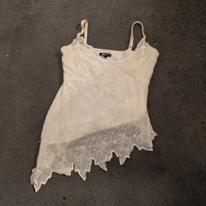 Vit spetsig linnetopp  - Säljer en vit linnetopp med tunna axelband och spetsdetaljer. Toppen har broderat blommönster.