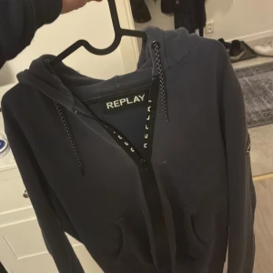 Ziphoodie från Replay - Snygg mörkblå zip hoodie från Replay med huva och dragkedja framtill. Jackan har coolt Replay-märke på ärmen och praktiska snören vid huvan. Perfekt för dig som gillar streetwear och vill ha en stilren look.