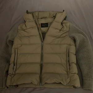Pufferjacka från Massimo Dutti - Snygg pufferjacka från Massimo Dutti med stickade ärmar i matchande ton. Jackan har huva, dragkedja framtill och quiltad kropp. Perfekt för dig som gillar en chill och modern look med extra komfort under höst och vinter. Endast använd en vinter. Kom med bud!