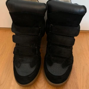 Svarta höga sneakers med kardborre - Säljer ett par svarta höga sneakers med platt sula och rund tå. Skorna har flera breda kardborreband istället för snörning, och är tillverkade i mocka och skinn med beige insida. Yttersulan är i gummi med brun ton. Perfekt för dig som gillar chunky och sportig stil.