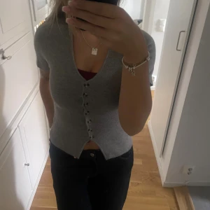 Grå v-ringad topp med knappar - Säljer en grå v-ringad topp med korta ärmar och snygga knappar framtill. Modellen är figurnära och har en croppad passform som ger en modern look. Perfekt att styla med jeans eller kjol för en chill vibe.