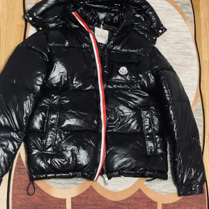 Svart Moncler Montbeliard  - Svart glansig dunjacka från Moncler med huva och klassisk logga på bröstet. Jackan har röda och vita detaljer längs dragkedjan, flera fickor med dragkedja och quiltad design. Perfekt för kalla dagar och riktigt snygg streetstil. Storlek L passar M med 
