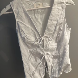 Vit linneväst från Gina Tricot - Snygg vit linneväst från Gina Tricot med omlott-design och två knytband framtill. Västen har v-ringad hals och är tillverkad i mjuk bomull. Använd en gång. Nypris 359kr
