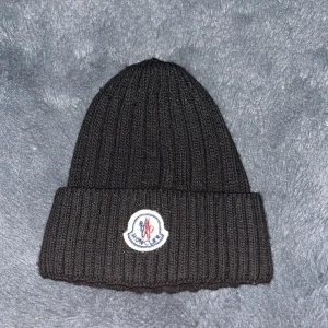 Svart ribbad Moncler mössa - Svart ribbstickad mössa från Moncler med klassisk uppvikt kant och broderad logotyp framtill. Mössan är tillverkad i mjuk bomull och har en clean, stilren look som passar perfekt till streetwear. Snygg detalj med Moncler-märket i vitt, blått och rött.