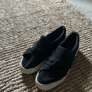 Svarta slip-on sneakers från Anna Field - Svarta slip-on sneakers från Anna Field med vit sula och snygg knutdetalj framtill. Skorna är i textil och har en rund tå samt diskreta prickar på innersulan. Perfekta för en chill och stilren look.