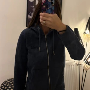 Mörkblå hoodie från Superdry - Mysig mörkblå hoodie från Superdry med dragkedja, huva och snörning. Tröjan har broderad logga på bröstet och mjukt foder på insidan. Perfekt för kyliga dagar och chill vibes.