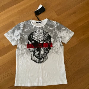 Vit och grå t-shirt med döskalle Iconic (barn) - Snygg t-shirt från Iconic i vitt och grått med ett stort döskalletryck framtill och en röd detalj över ögonen. T-shirten har rund halsringning och korta ärmar. Bröst 47 Längd 65. Ca 122-128. 