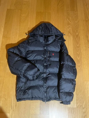 Svart pufferjacka från Polo Ralph Lauren - Svart pufferjacka från Polo Ralph Lauren med huva, dragkedja och en röd broderad logga på bröstet. Jackan har flera fickor med dragkedja . Perfekt för kalla dagar och har en riktigt schysst streetstil. En knapp saknas som man ser på bilden 