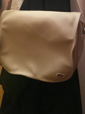 Beige axelväska från Lacoste - Säljer en stilren beige axelväska från Lacoste med klassisk krokodillogga på framsidan. Väskan har ett justerbart axelband och insidan är fodrad med randigt tyg och har en praktisk dragkedja. Perfekt för dig som gillar enkel och snygg design.