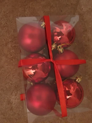 Julkulor - Denna bild visar en förpackning med sex röda julgranskulor, både blanka och matta, med guldfästen. Kulorna är snyggt förpackade i en transparent ask med röd rosett. Perfekt för att dekorera granen inför julen. Inga synliga defekter eller slitage.