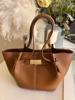 Brun handväska med gulddetalj - Snygg liten brun handväska i skinnimitation med två handtag och dekorativ guldfärgad låsdetalj framtill. Väskan har en mjuk, rundad form och fina sömmar samt knyt på sidorna för extra stilpoäng. Perfekt storlek för det viktigaste och passar till många looks.