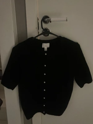 Svart kofta från H&M med knappar - Svart kofta från H&M i storlek XS. Koftan har korta ärmar och är försedd med silvriga knappar längs hela framsidan. Perfekt att slänga över en topp för en chill look. Materialet känns mjukt och skönt mot huden. 