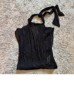 Svart omlottopp med knyt från Zara - Säljer en svart omlottopp från Zara. Toppen har ett snyggt knytband vid halsen och är gjord i ett lätt, transparent material med subtila glittriga ränder. Aldrig använd 