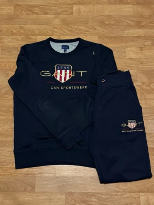 Mörkblå tracksuit från GANT - Säljer ett par mörkblå mjukisbyxor från GANT med broderad logga och text på benet. Byxorna har snörning i midjan och är gjorda i mjukt bomullsmaterial. Perfekta för chill eller sportiga dagar.