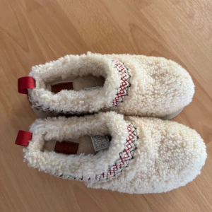 Vita fluffiga slip-ins från UGG tazz 37 - Mysiga slip-in skor från UGG i krämvit färg med fluffigt fårskinnsmaterial. Dekorativt broderi i rött och svart runt öppningen samt röd hälflik. Sulan är robust och mönstrad för bra grepp. Perfekta för kalla dagar när du vill ha det extra bekvämt.
