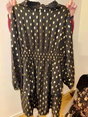 Vacker svart klänning med guldprickar - Supersnygg svart långklänning med glittriga guldprickar och långärmad design. Klänningen har resår i midjan för en skön passform och volangdetaljer nedtill. Perfekt för dig som vill sticka ut med ett coolt mönster och metallic-vibe. Väldigt vacker