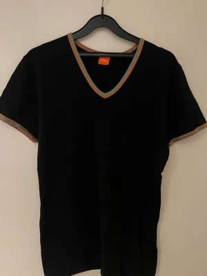 Svart t shirt  med färgglada kanter från BOSS - Svart t shirt  från BOSS med v-ringad hals och korta ärmar. Kanterna vid halsringning och ärmslut har färgglada ränder. T shirtsen material känns mjukt och har en klassisk passform som funkar till många stilar.