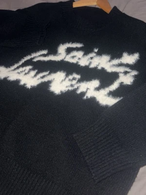 Svart stickad Saint Laurent tröja - Säljer en svart stickad tröja från Saint Laurent med vit, fluffig logga framtill. Tröjan har rund halsringning och långa ärmar med breda ribbade muddar. Perfekt för dig som vill ha en statement piece i garderoben.