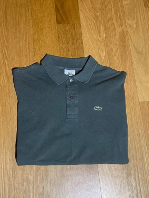 Mörkgrön pikétröja från Lacoste - Säljer en stilren mörkgrå pikétröja från Lacoste med klassisk krage och två knappar framtill. Tröjan har korta ärmar och det ikoniska gröna krokodilmärket broderat på bröstet. Materialet är bomull och modellen har en normal passform. Storlek 4 = S