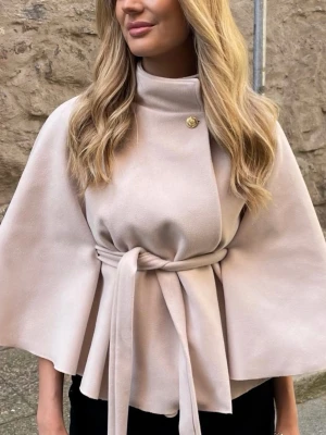 Beige poncho - Säljer en elegant beige poncho med capeformade ärmar och högt krage. Jackan har ett snyggt bälte i midjan och en dekorativ knapp vid halsen. Perfekt för dig som gillar stilrena och trendiga plagg med en lyxig känsla. Endast använd en gång