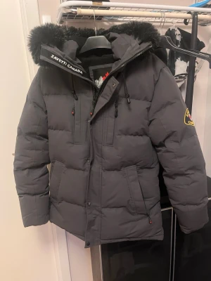 Mörkgrå pufferjacka från Zavetti Canada - Säljer en mörkgrå pufferjacka från Zavetti Canada med fluffig svart päls på huvan och zavetti canada logga på ärmen. Jackan har dragkedja, knappar och flera fickor framtill. Perfekt för kalla dagar och har en riktigt schysst   streetstil. ,   säljs pågrund av dene för stor för mig . Den är perfekt skick , har använts men inga hål eller något . Tyget man hänger jackan har gått sönder men skapar inga problem för användning . 