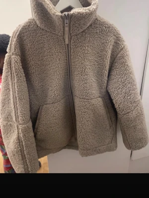 Beige teddyjacka med dragkedja - Mysig beige teddyjacka med hög krage och två stora fickor framtill. Jackan har en rak och loose passform, långärmade ärmar och stängs med en rejäl dragkedja. Perfekt för kalla dagar när du vill vara både varm och snygg. Pris kan diskuteras 