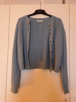 Ljusblå stickad kofta från Mango (M) - Säljer en ljusblå stickad kofta från Mango med snyggt kabelmönster och ribbade muddar. Koftan har rund hals, långa ärmar och knappar framtill i en neutral ton. Perfekt att slänga över en t-shirt för en chill look. Den är otroligt mjuk och även stretchig. Storlek M, använd få gånger 