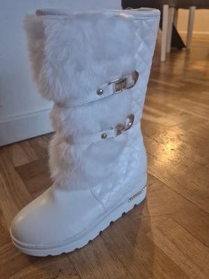 Vita quiltade boots med päls och spännen - Säljer ett par vita boots med quiltad design, fluffig päls på skaftet och snygga guldfärgade spännen. Insidan är fodrad med mjuk syntetpäls för extra värme. Sulan är platt och har dekorativa detaljer i guld. Perfekta för vinter och kalla dagar!