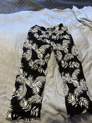 Svarta byxor med vit palmprint från Lindex - Säljer ett par svarta byxor från Lindex med coolt vitt palmbladsmönster. Byxorna har rak passform och är gjorda i ett lätt och skönt material, perfekta för chill dagar. Storlek 146, passar dig som är 10-11 år.