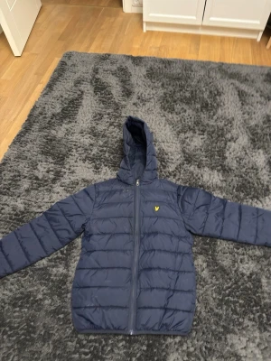 Blå pufferjacka från Lyle & Scott 10/11 år - Säljer en blå pufferjacka från Lyle & Scott Junior med huva och dragkedja framtill. Passar dig som är runt 146-152. Behöver en liten tvätt bara.